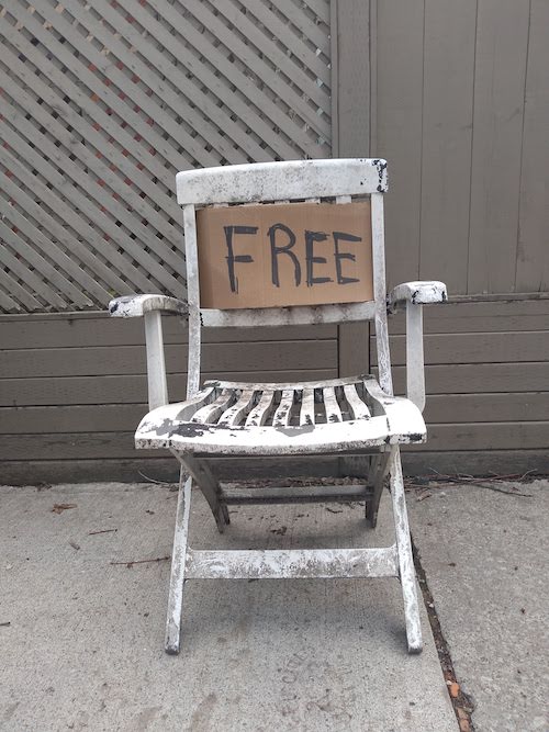 Image of a battered old plastic chair sitting on the sidewalk with a handwritten cardboard sign on it saying FREE.