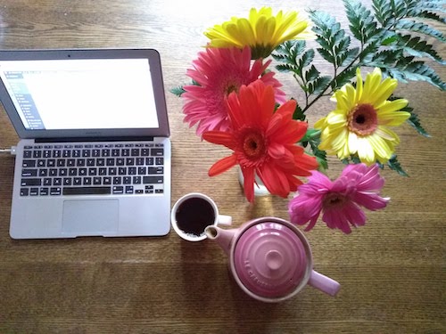 A desktop with a laptop computer, bouquet of daisies, and a pink teapot.